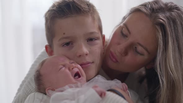 Teenage Boy Looking at Camera Standing Indoors with Crying Baby Sister and Beautiful Mother