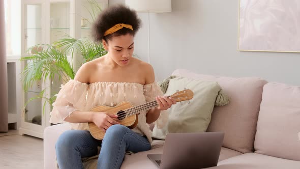 Young african american woman taking online ukulele music guitar lessons tutorial on laptop