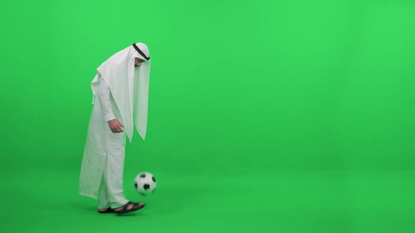 An Arab Man in a White Robe on the Background of a Green Background Plays with a Soccer Ball Kicks