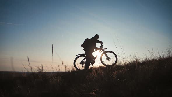 The Silhouette of the Mountain Bicycle Rider on the Hill with Bike at Sunset. Sport, Travel and