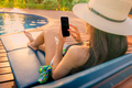 Asian woman with hat and swimsuit sitting in chair at poolside and using smartphone on vacation. - PhotoDune Item for Sale