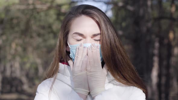 Close-up of Young Brunette Woman in Protective Mask and Gloves Coughing in Sunny Spring Park
