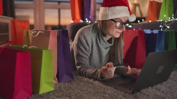 Woman with Glasses Wearing a Santa Claus Hat is Lying on the Carpet and Makes an Online Purchase