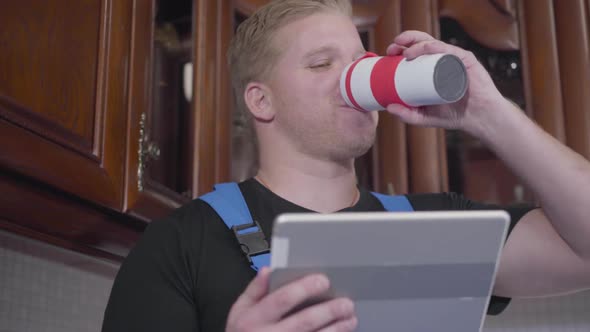 Close-up of Young Caucasian Redhead Man Sitting with Tablet and Drinking Tea or Coffee. Unscrupulous