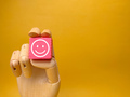 Wooden hand holding pink cube with smile icon on yellow background. Business concept. - PhotoDune Item for Sale