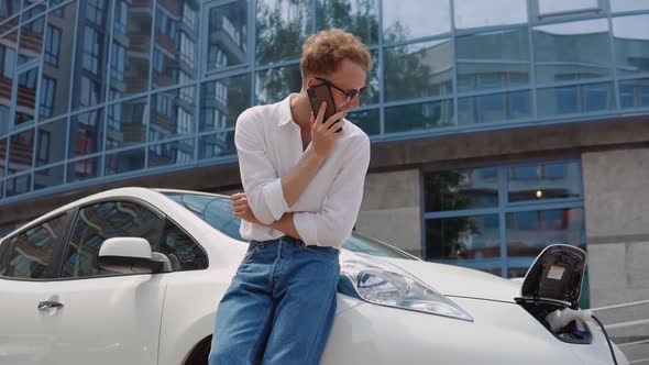 Stylish Modern Young Curly Man Charges His Electric Car in the Yard of a Residential Complex and