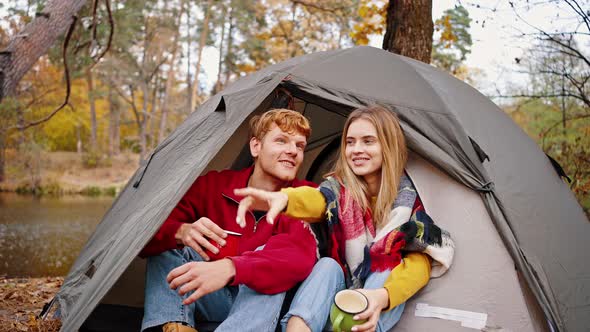 Young Couple Laughing and Pointing Forefingers Holding Cups of Tea Sitting in Tent Near River at