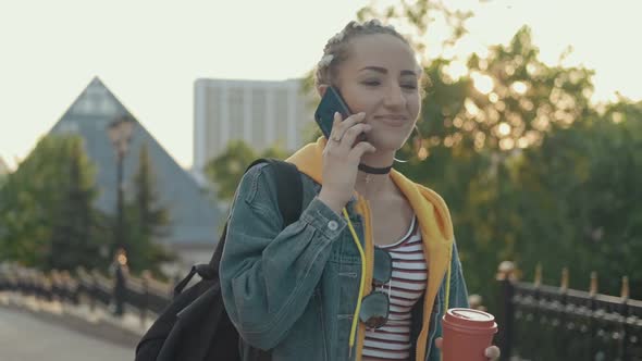 Joyful Hipster Girl Using Cellphone Outdoors with Cup of Coffee or Tea in Hand