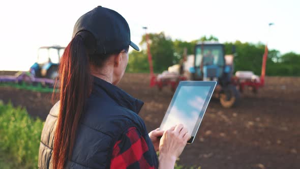 Woman Uses Specialized App on a Digital Tablet PC on Background of Working Tractor in Field
