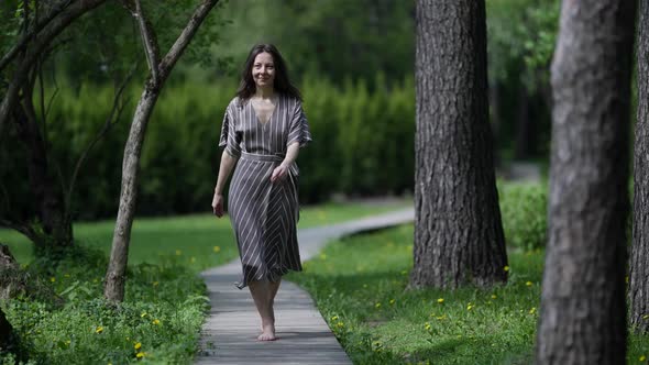 a Brunette in a Striped Dress and Barefoot Merrily Walks Along a Wooden Path in the Park