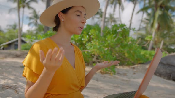 Smiling Pretty Girl in a Hat and Yellow Dress Having Online Call Using Laptop on the Tropical Beach