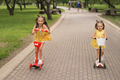 Funny laughing girls ride scooters on paths in the park. - PhotoDune Item for Sale