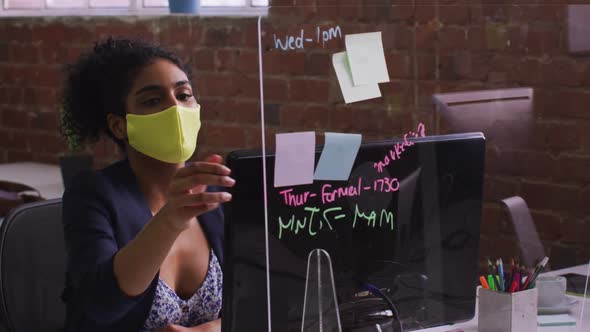 Mixed race businesswoman using memo notes sitting in front of computer wearing face mask