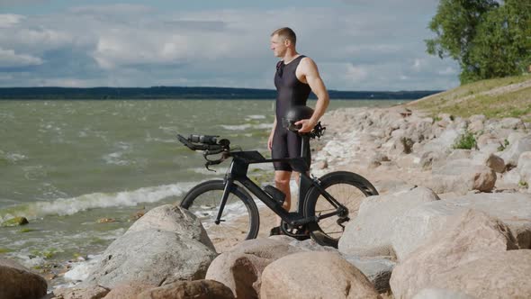 Handheld Pro Triathlete Stand Near the Lake Holding a Bicycle Helmet in His Hands a Cyclist in