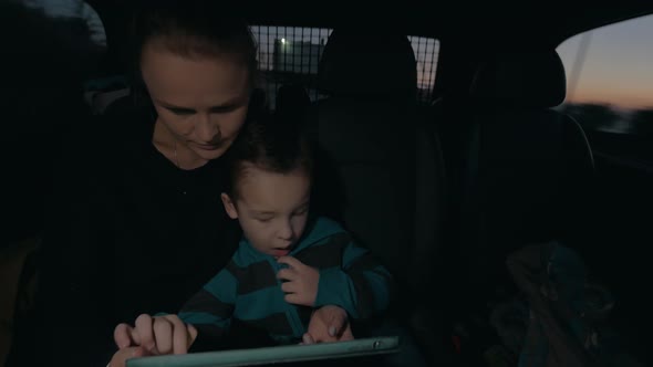 View of woman with son in the dark riding in car on back seat, watching computer tablet