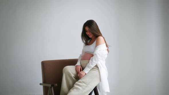 Pregnant Girl Sits on a Chair Against a White Background and Poses for the Camera