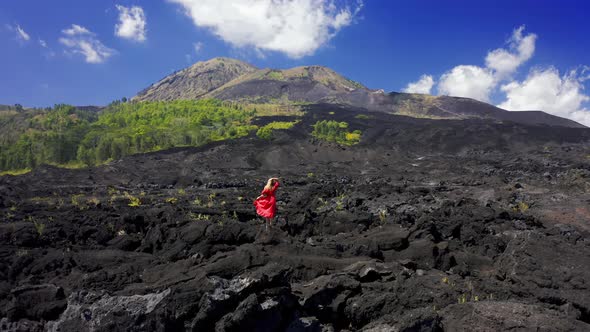 Young Woman in Red Dress on Frozen Black Lava and Green Trees on the Slope of Volcano Batur on Bali