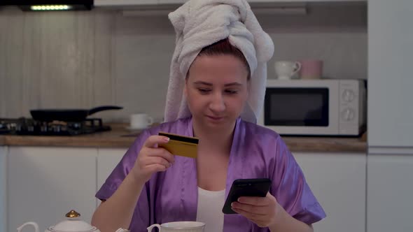 Young Woman is Sitting at Table in Kitchen Makes an Online Purchase Using Smartphone and Credit Card