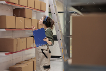 etails, listening music while analyzing clipboard with products checklist in storehouse. Employee working at merchandise delivery in warehouse