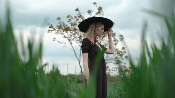 Blonde Holds Big Black Hat With Her Hand On Her Head.