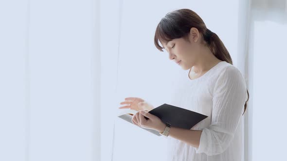 Beautiful young asian woman standing reading book for exam with serious and stress in the bedroom.