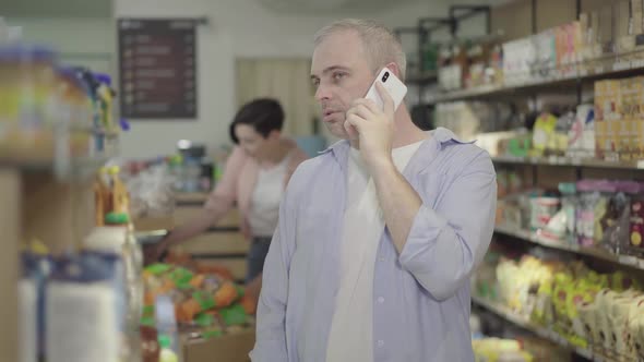 Serious Caucasian Man Talking on the Phone in Grocery. Concentrated Husband Consulting Wife As