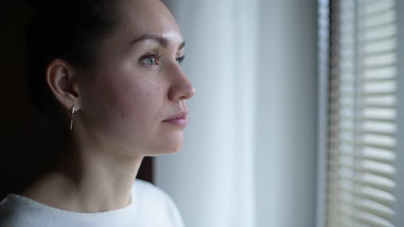 Girl looks thoughtfully out the window at the evening street through the blinds