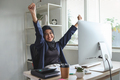 Muslim business woman with arms raised sitting on chair at office desk celebrating achievement - PhotoDune Item for Sale