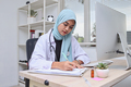 Muslim woman doctor writing on clipboard while using laptop on desk in hospital. - PhotoDune Item for Sale