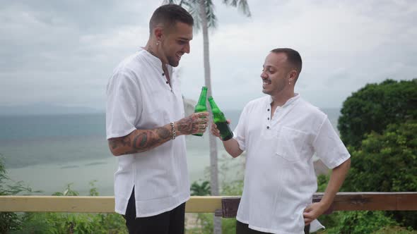 Cheerful Male Gay Couple Clinking Bottles of Beer While Standing a Tropical Island Terrace with a