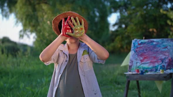 Artistic Child Cute Boy in Hat Shows His Palms in Paint While Drawing on in Park on a Summer Evening