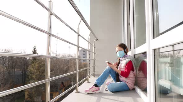 Teenager Girl in Mask and Headphones, Listening To Music on Smartphone, on Open Balcony. Spring