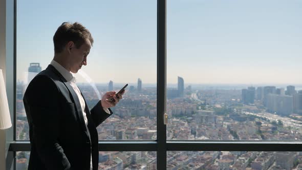 Successful Businessman Is Happy About Getting Great News on Smartphone By the Window in Office