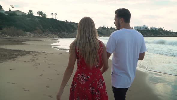 Bak View Young Couple Holding Hands Walking Towards Sunset on Empty Beach Speaking with Each Other