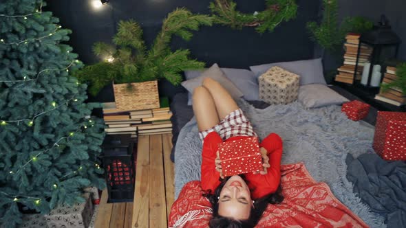 Pretty girl in cozy bedroom at Christmas. Beautiful young woman lying on bed with a gift in New year