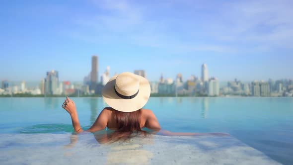 Lonesome Woman With Floppy Hat in Rooftop Infinity Pool With View of Urban Sunny Cityscape, Back Vie