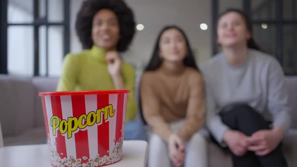 Closeup of Popcorn Bucket with Three Blurred Young Multinational Women Watching TV at Background