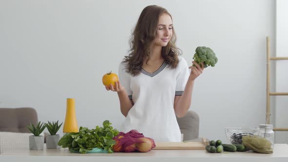 Portrait Young Woman Holding Yellow Tomato and Broccoli Standing at the Table in Modern Kitchen