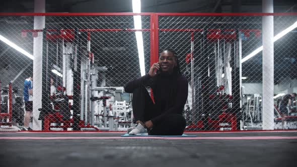 Smiling Africanamerican Woman Sitting on Yoga Mat in a Gym and Talking with Someone on Her Phone