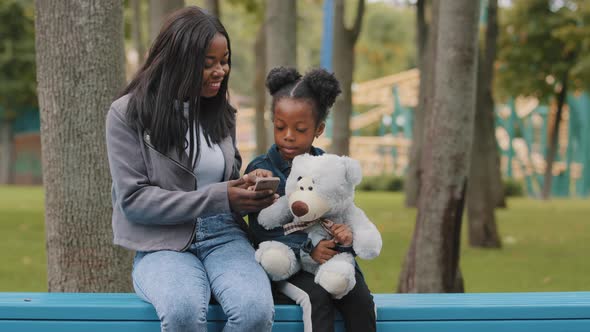 Positive Mom and Daughter Sitting on Bench in Park Mother with Child Smiling Looking at Phone