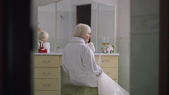 Reflection of Sad Senior Greyhaired Woman Sitting at Bathroom Nightstand with Wedding Dress