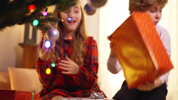 Excited Little Boy and Girl at New Year Tree Indoors Checking Gifts Smiling