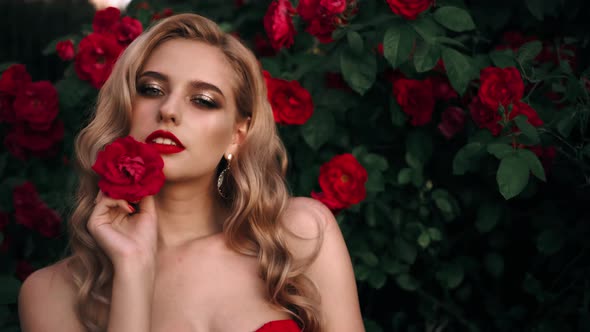 Fabulous Girl with Red Lips in Red Dress on Awesome Summer Roses Background. Fantasy Woman Portrait