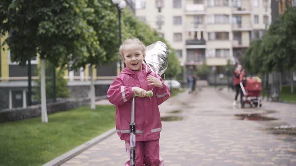 Portrait of Cheerful Happy Girl Riding Scooter Outdoors. Laughing Cute Caucasian Child Holding