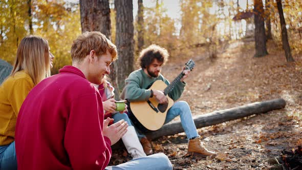 Friends Playing Guitar and Clapping Hands Listening to Music Sitting on Logs in Autumn Wood