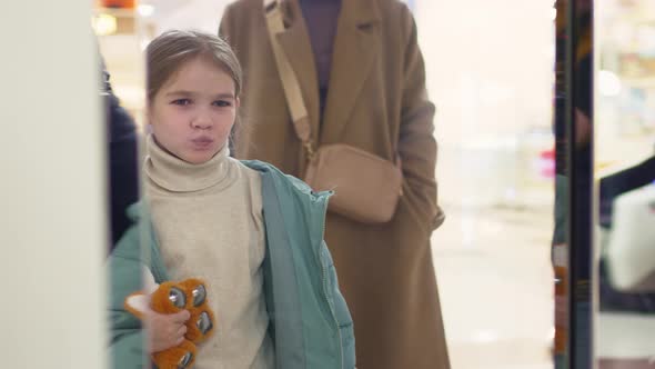 Funny Little Girl with Toy Writhes Faces in Door Mirrored Elevator in Mall