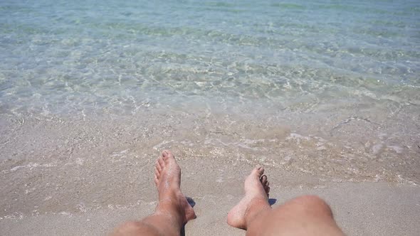 Closeup of Men's Feet Lying on the Seashore Small Waves Hitting Their Feet on the Shore in Slow