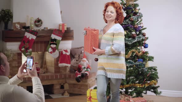 Husband Taking Photos of Cheerful Caucasian Wife Posing with Christmas Gift with New Year Tree at