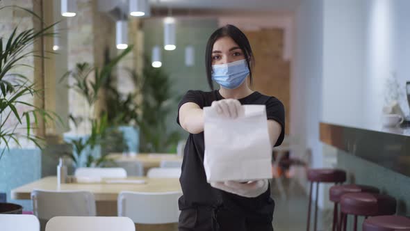 Front View Young Woman in Covid Face Mask Stretching White Disposable Paper Bag with Takeaway Food