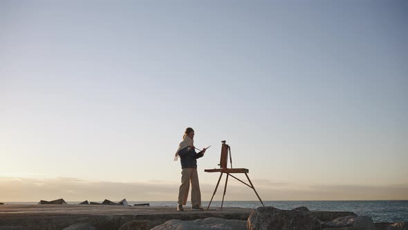 Woman Painter Drawing Outdoors Standing with Easel on Sea Bank in Evening Over Colorful Sunset Sky